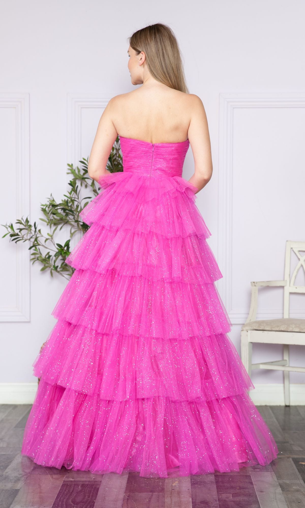 A woman faces away from the camera in a Strapless Long Tiered Glitter Prom Ball Gown 9386, featuring bright pink tulle, in a light-walled room with a plant and a white bench.